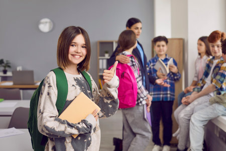 Portrait of happy girl school student showing thumb up. Beautiful smiling schoolgirl wearing casual clothes standing with backpack on background of her classmates in classroom and looking at cameraの写真素材