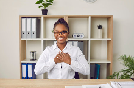 Happy African woman in white shirt and glasses sitting at office table at business meeting or Internet conference, holding hands at heart, expressing gratitude, saying thank you for this opportunityの写真素材