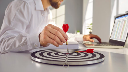 Skillful confident business man works on laptop, sets goal and hits mark as he points red arrow sharp in bulls eye of round archery shooting target circle on desk in office workplace. Objects close upの写真素材