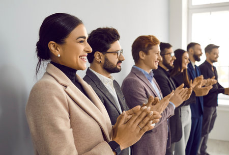 Happy smiling professional young business people clap their hands. Selective focus on attractive men and women lined up along the wall. Side view of stylishly dressed colleagues near the window.の写真素材