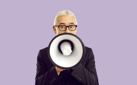 Mature businessman with loudspeaker isolated on purple background scream make announcement. Old man in glasses hold megaphone talk speak to public. Attention concept.の写真素材