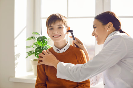Happy school boy getting an ear check up at the clinic. Woman doctor doing an ENT examination to a child patient. Professional otolaryngologist uses an otoscope to examine ears of a little smiling kidの写真素材