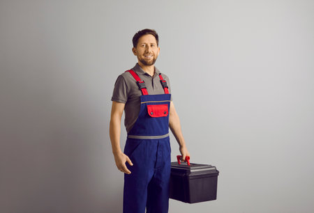 Portrait of a happy smiling confident repairman with beard holding a tool box in hands isolated on gray wall background. Professional young handyman worker standing cheerful in blue overall.の写真素材