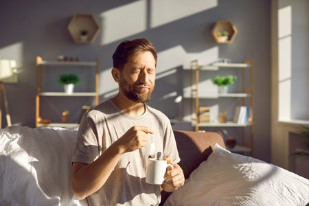 Man drinks tea as he wakes up in the morning. Young man sitting on the sofa in a sunny room at home, holding a cup and a tea bag, drinking tea and squinting in the morning sunの写真素材