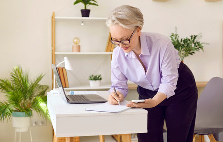 Senior business woman making notes in notebook while looking at mobile phone screen. Successful stylish mature woman at her workplace in bright modern office. Business and senior people concept.の写真素材