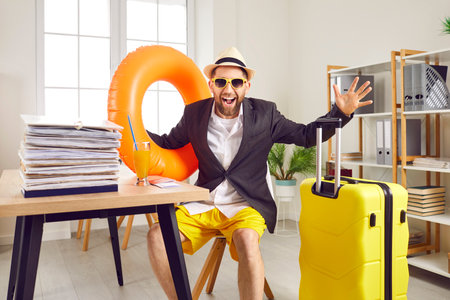 Happy funny excited man in suit jacket, shirt, sun hat, summer shorts and sunglasses sitting at office desk with paperwork, beach ring, orange juice and suitcase. Holiday vacation annual leave conceptの写真素材