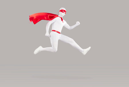 Funny super hero in a red cape, eye mask and white bodysuit flying in the air on a gray color background. Anonymous man in a superhero costume hurrying to perform his duty of saving the worldの写真素材