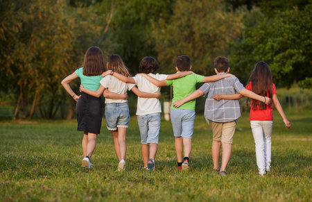 View from back boys and girls walk together on grass hugging each other walking in casual summer...