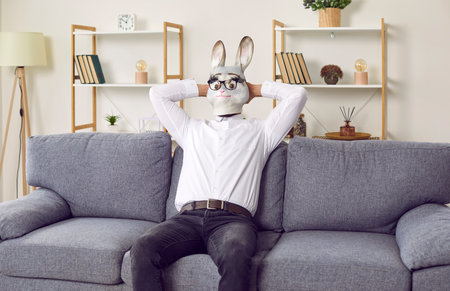 Business animal man enjoying weekend. Funny young guy in rabbit face mask, white shirt and glasses sitting alone on comfortable sofa at home, with hands behind head, relaxing and enjoying leisure timeの写真素材