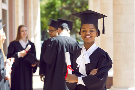 Young smiling african american girl student in a university graduate gown and diploma in her hands. Woman with crossed arms outdoor on the background of classmates. Graduation and education concept.の写真素材