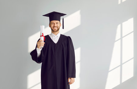 Young smiling joyful man student in university graduate gown on a grey background. Happy guy holding diploma in his hands indoors and proudly looking at camera. Graduation and education concept.の写真素材