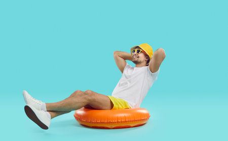 Happy satisfied young man enjoys summer sea vacation sitting on inflatable swimming circle. Joyful guy in summer outfit sits on inflatable circle with his hands behind his head on blue backgroundの写真素材