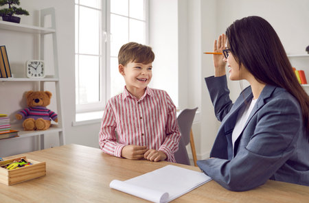Friendly school counselor or psychologist communicating with a child. Young woman sitting at her desk with a clipboard and talking to a happy, cheerful little boy. Childrens psychology conceptの写真素材