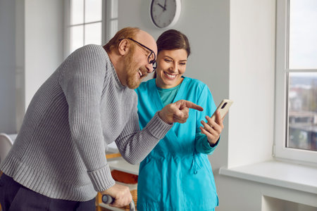 Red-haired, elderly bearded man with caring nurse are laughing and having fun in a nursing home. Pensioner dressed in gray warm sweater, pointing at mobile phone screen. Taking care of elderly people.の写真素材