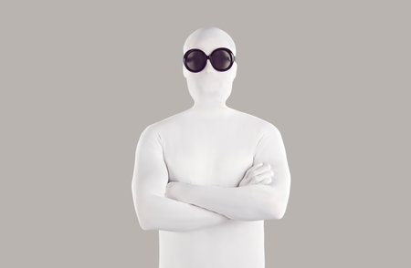 Portrait of person in bodysuit disguise and sunglasses. Man with secret, hidden personality, wearing white spandex costume and black round glasses standing with folded arms on grey color backgroundの写真素材