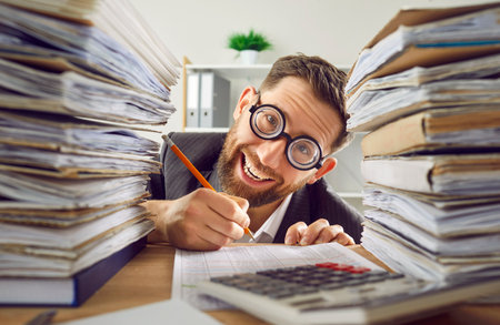 Funny bookkeeper has much paperwork. Crazy man in suit and round glasses sitting at office desk, working with files, writing something with pencil and smilingの写真素材