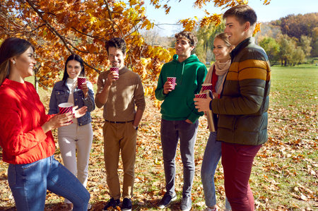 Group of a young friends drinking coffee or tea in the autumn park. Happy caucasian people girls and boys talking to each other outdoors. Smiling students chatting and communicate together.の写真素材