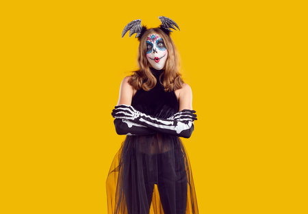 Studio portrait of child in Halloween costume. Teenage girl with sugar skull make up standing with arms folded isolated on yellow background and looking at camera with funny surprised face expressionの写真素材