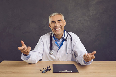 Portrait of cheerful physician at office table. Happy senior male doctor in white lab coat with stethoscope sitting at desk, looking at camera, gesturing, smiling and welcoming you at consultationの写真素材
