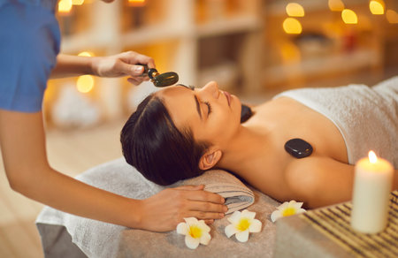 Young woman enjoying face massage at spa salon. Specialist doing facial massage with green jade roller to beautiful relaxed woman lying on bed on towel with flowers. Beauty, skin care conceptの写真素材