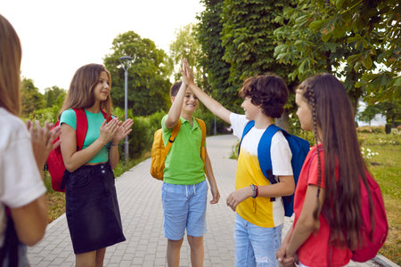 Joyful little friends boys and girls in casual clothes with kids backpacks on their backs posing for childrens portrait in summer park high five to each other and saying goodbye after schoolの写真素材