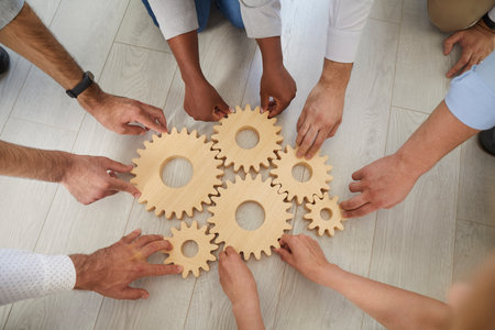 Team of diverse business people joining gear wheels. Group of multiracial men and women connecting wooden cog wheels on the floor. Cropped shot, human hands, from above. Teamwork, cooperation conceptsの写真素材