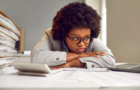 Stuck in this office. Tired, bored, frustrated young African American woman employee in glasses sitting at office desk with paperwork workload and waiting for end of work day with sad face expressionの写真素材