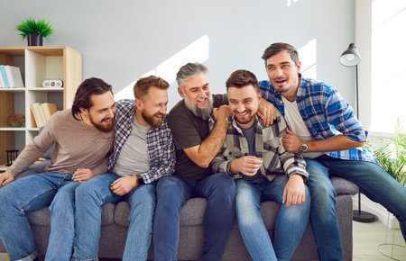 Portrait of a male bearded friends sitting on sofa, smiling and having fun at home in the living room. Photo of group of brutal friendly men in casual clothes hugging indoors on the party.の写真素材