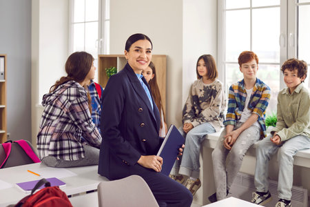 Smiling female teacher standing with her students in classroom. Confident junior boys and girls...