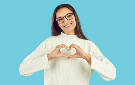 Portrait of beautiful friendly woman who shows loving gesture and expresses her sympathy to you. Happy smiling Caucasian woman showing heart folded with fingers, isolated on light blue background.の写真素材