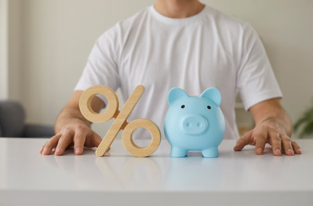 Cropped shot of a young man sitting at a white desk with a wooden percent symbol and a blue ceramic piggy bank. Closeup shot, close up view. Money, finance, percentage rate, bank interest conceptの写真素材