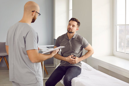 Young sick man sitting on the couch in the doctors office and pointing to stomach during medical...