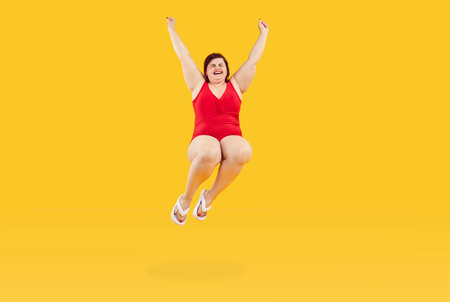 Happy funny fat woman in red swimsuit and flip flops jumping isolated on yellow color background. Joyful plus size lady enjoying summer holiday, having fun and jumping in swimming pool or ocean waterの写真素材