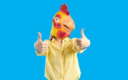 Studio shot of young girl with funny bird face showing thumbs up with both hands. Woman wearing...