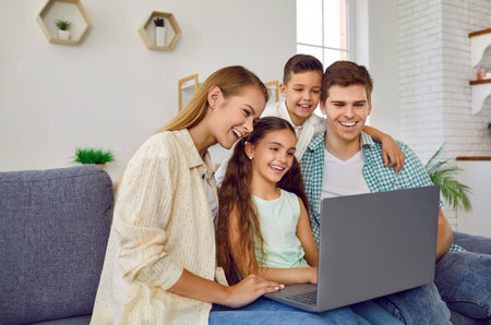 Parents together with their children enjoy weekend and laugh watching videos on computer at home. Happy young Caucasian family sitting on sofa in living room and looking at laptop screen.の写真素材