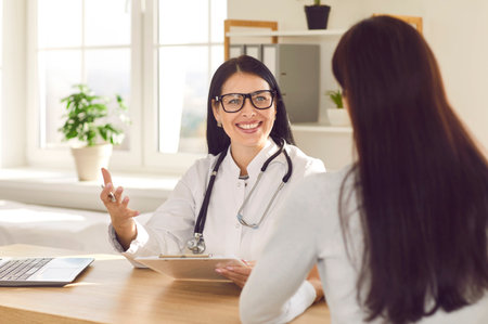Portrait of smiling doctor sitting in office wearing stethoscope and holding report file with appointment and giving consultation to a woman sitting back during medical examination in clinic.の写真素材