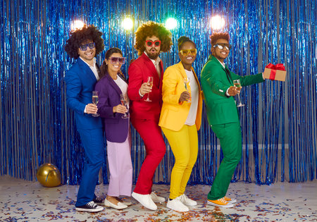 Friends having fun at New Years party in colorful costumes and with champagne in their hands. Cool multiracial young people stand in row against shiny foil curtain during retro themed party.の写真素材