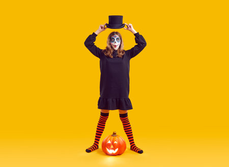 Portrait of funny teenage girl with Halloween makeup looking surprised on orange background in studio. Child in black dress and striped leggings holds hat over his head while standing next to pumpkin.の写真素材