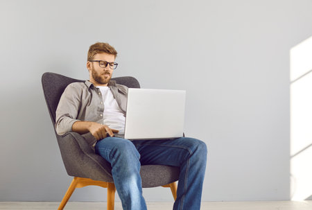Portrait of a young bearded businessman sitting on chair in empty space with laptop working or studying online isolated on grey wall background. Remote work and freelance career concept.の写真素材