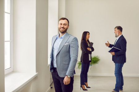 Portrait of happy smiling man looking at camera with realtor showing and discussing new apartment with his wife on background. Successful family ready to become homeowners. Real estate market conceptの写真素材