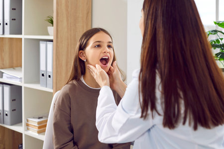 Child patient visits doctor. Woman pediatrician or otolaryngologist asks kid to open mouth and looks at sore throat. Endocrinologist examines little girl, holds her neck and checks her thyroid glandsの写真素材