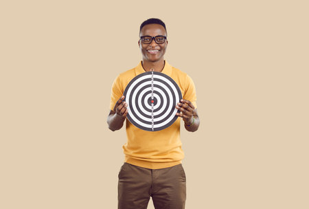 Cheerful ethnic man holding shooting target. Studio portrait of happy smiling handsome young African American guy in casual T shirt holding dart board with concentric circles. Setting goals conceptの写真素材