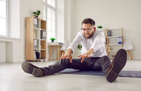 Funny business man having fitness workout in office, sitting on yoga mat on floor and trying to do...