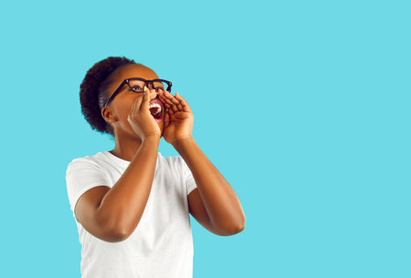 Young woman in white T shirt and glasses puts hands at mouth and shouts loudly on blank blue text copy space background. Loud Afro American girl advertises something and shares important informationの写真素材