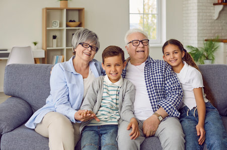 Portrait of a happy grandparents sitting on sofa with their grandchildren boy and girl in the living room at home looking at camera and smiling. Kids enjoying leisure time with grandma and grandpa.の写真素材
