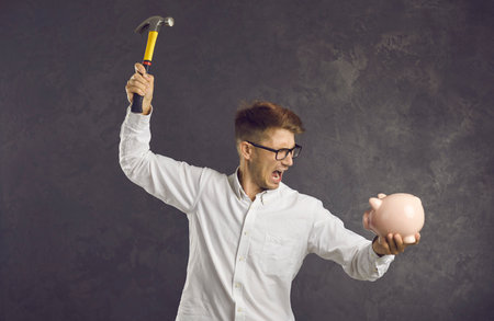Financial difficulties. Emotional and crazy frustrated young man is going to break piggy bank with hammer. Angry man shouting holding piggy bank and hammer in his hands standing on gray background.の写真素材