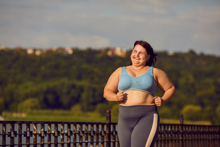 Happy overweight woman jogging. Cheerful smiling motivated beautiful fat large plump chubby stout lady in sports bra running on bridge over river on sunny summer morning with green trees in backgroundの写真素材