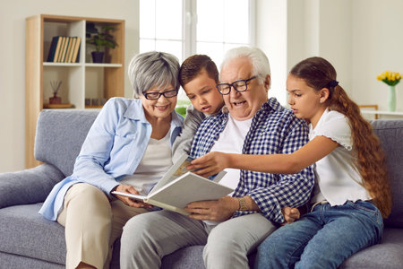 Portrait of a happy grandparents reading a book to their grandchildren or looking through a family photo album sitting on sofa at home. Kids enjoying leisure time with grandma and grandpa on weekendの写真素材