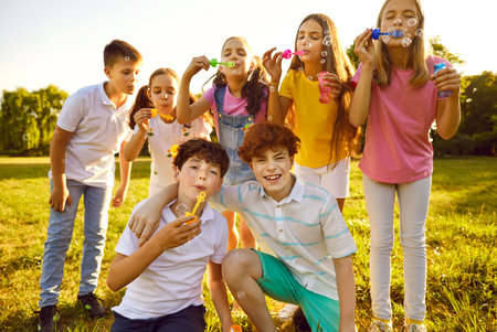 Portrait of happy kids friends standing on green grass in park blowing bubbles together. Group of...