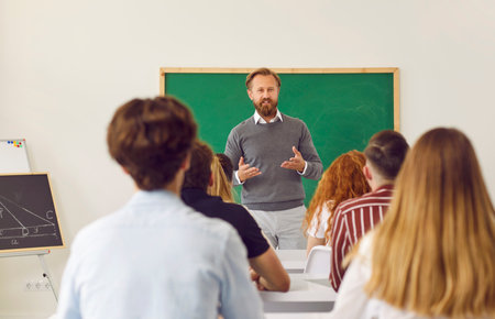 Man teaching group of young people. Math teacher talking to pupils in class. Adult man in shirt and jumper standing in front of blackboard in classroom and giving lecture to school or college studentsの写真素材